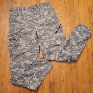 Pode incluir: Calças cargo de camuflagem digital em tons de cinza, bege e verde. As calças apresentam vários bolsos e fecho de botão. O tecido tem um padrão pixelizado, típico de roupas de estilo militar.