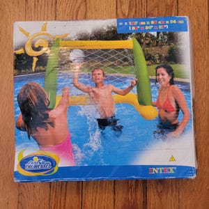 Peut inclure: Un panier de basket-ball gonflable pour piscine jaune et vert avec un filet. La boîte montre trois personnes jouant dans une piscine avec le panier. La boîte indique "The Wet Set" et "INTEX". Les dimensions sont de 127 cm x 86 cm x 94 cm.