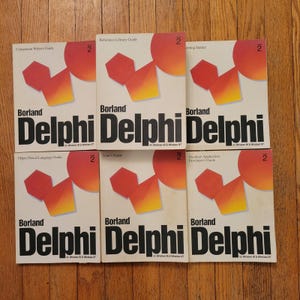 Puede incluir: Una colección de seis manuales de software Borland Delphi. Cada libro tiene una cubierta blanca con el título "Delphi" en letras negras grandes. Las cubiertas presentan un diseño geométrico rojo, naranja y amarillo y el número "2".