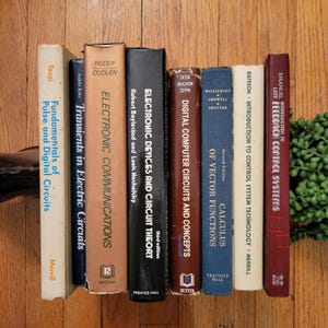 May include: A collection of vintage books with titles related to electronics, circuits, and calculus. The books are arranged vertically on a wooden surface, with visible titles like "Electronic Communications" and "Digital Computer Circuits and Concepts."