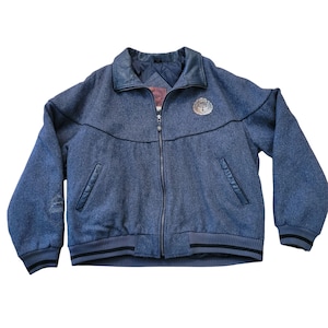 May include: A blue wool bomber jacket with a black leather collar and a zip closure. The jacket has a logo patch on the left chest and two pockets on the front.