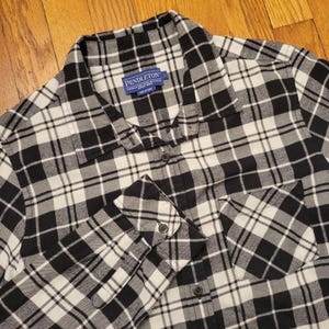 Könnte beinhalten: Ein schwarz-weiß-graues kariertes Button-Down-Hemd mit Kragen und Brusttasche. Das Hemd hat lange Ärmel mit geknöpften Manschetten. Das Markenetikett "Pendleton" ist am Kragen sichtbar.