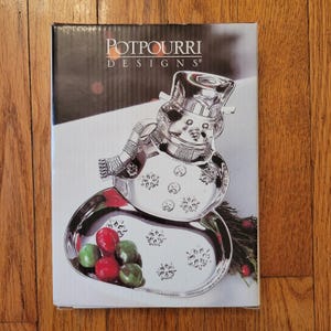 Puede incluir: Una fuente plateada con forma de muñeco de nieve con sombrero, bufanda y detalles de copos de nieve. El plato contiene caramelos rojos y verdes. La caja dice "POTPOURRI DESIGNS".