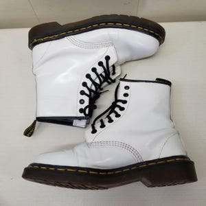 May include: Pair of white leather boots with black laces and a brown sole. The boots have a classic design with visible stitching and a pull tab at the back. The soles are approximately 1 inch thick.