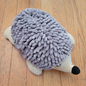 May include: A plush hedgehog toy featuring a textured gray back made of soft, chenille-like fabric loops. The underside, face, and limbs are crafted from smooth, cream-colored fabric, with a small black nose and dark eyes.