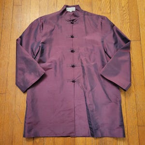 May include: A long-sleeved, plum-colored jacket with a mandarin collar and frog closures. The jacket has a silky sheen and a relaxed fit. The sleeves are three-quarter length. The garment is laid flat on a wooden surface.