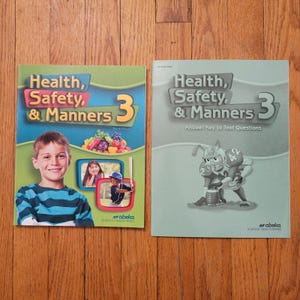 Puede incluir: Se muestran dos libros de texto titulados "Health, Safety, & Manners 3". Una portada presenta a un niño, fruta y dos fotos insertadas. El otro libro es de color verde claro con una ilustración de un jugador de fútbol americano. Ambos libros son de Abeka.