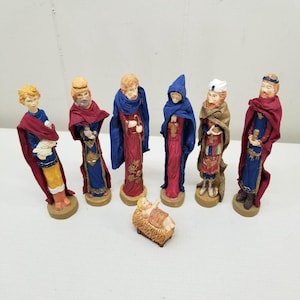 May include: A set of seven religious figurines. The figures are dressed in robes of red, blue, and gold. One figure is a baby in a manger. The figurines are on a light brown base.