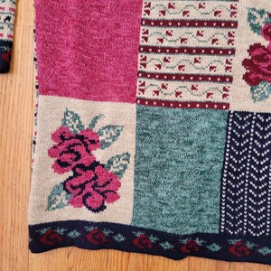 Op de afbeelding: Close-up van een patchwork trui met een bloemenontwerp. De trui heeft vierkanten in roze, teal en beige, met bloemen- en geometrische patronen. De onderste zoom heeft een donkerblauwe rand met rode roosdetails.