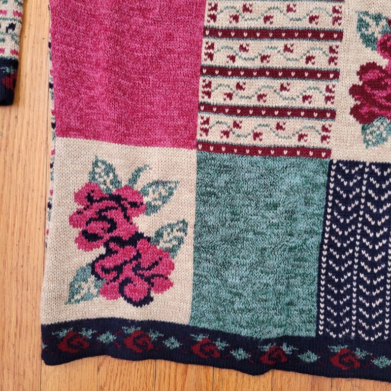 Op de afbeelding: Close-up van een patchwork trui met een bloemenontwerp. De trui heeft vierkanten in roze, teal en beige, met bloemen- en geometrische patronen. De onderste zoom heeft een donkerblauwe rand met rode roosdetails.