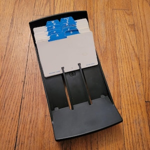 Könnte beinhalten: Ein schwarzer Rolodex-Kartenhalter mit weißen Karten und blauen alphabetischen Trennwänden. Die Datei ist geöffnet und zeigt die Karten und Trennwände. Der Rolodex befindet sich auf einer Holzoberfläche.