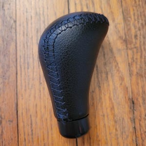 May include: Black leather gear stick knob with stitched detailing. The knob has a rounded top and a smooth, ergonomic design. The stitching is visible along the side of the knob. The base of the knob is black and glossy.