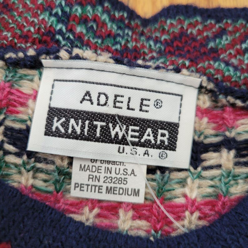 Op de afbeelding: Close-up van een label van een gebreide trui. Het label luidt "ADELE KNITWEAR U.S.A." en "PETITE MEDIUM". De trui heeft een kleurrijk patroon met rode, groene, blauwe en cr&egrave;mekleuren.