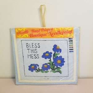 May include: A Bucilla Hand Painted Boutique Needlepoint kit featuring a white canvas with a blue flower design and the text "Bless This Mess".