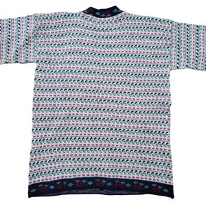 Op de afbeelding: Een vintage gebreide trui met een herhalend patroon van kleine rode, teal en marineblauwe vormen op een cr&egrave;mekleurige achtergrond. De trui heeft een marineblauwe kraag en zoom met een decoratief patroon. De mouwen zijn kort en de algehele stijl is casual.