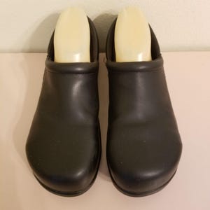May include: A pair of black leather clogs with a rounded toe and a padded collar around the opening. Each shoe contains a light beige plastic shoe tree insert to maintain the shape of the footwear. The exterior surface is smooth and matte.