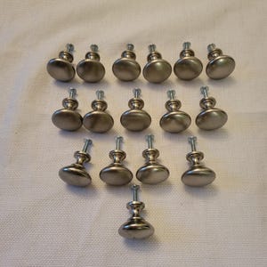 May include: A collection of sixteen silver-toned metal cabinet knobs arranged in rows. Each knob features a smooth, rounded top surface and a cylindrical base with an attached threaded metal screw extending from the bottom for installation purposes.
