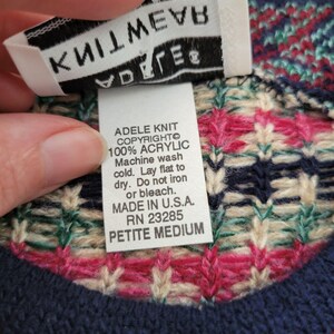 Op de afbeelding: Close-up van een gebreide trui met een label met de tekst "ADELE KNIT COPYRIGHTO 100% ACRYLIC Machine wash cold. Lay flat to dry. Do not iron or bleach. MADE IN U.S.A. RN 23285 PETITE MEDIUM." De trui heeft een marineblauw, roze en cr&egrave;mekleurig patroon.
