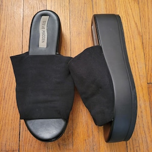 May include: A pair of black platform sandals with a slip-on design. The sandals feature a thick sole and a fabric strap across the top. The brand name "STEVE MADDEN" is visible on the insole.