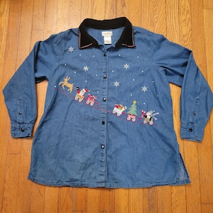 May include: A blue denim shirt with a black velvet collar and long sleeves. The shirt is decorated with embroidered Christmas motifs, including a reindeer, Santa Claus, and a Christmas tree. White snowflake embroidery adds detail to the shirt.