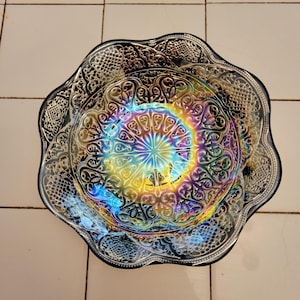 May include: An iridescent glass bowl with a scalloped edge. The bowl features intricate embossed patterns and a vibrant, rainbow-like sheen. The center of the bowl has a starburst design, with colors shifting from yellow to blue and purple.