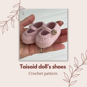 Può includere: Un paio di scarpine per bambole in crochet rosa con un bottone marrone su ogni scarpa. Le scarpine sono tenute in una mano. Il testo "Taisoid doll's shoes Crochet pattern" è sotto l'immagine.