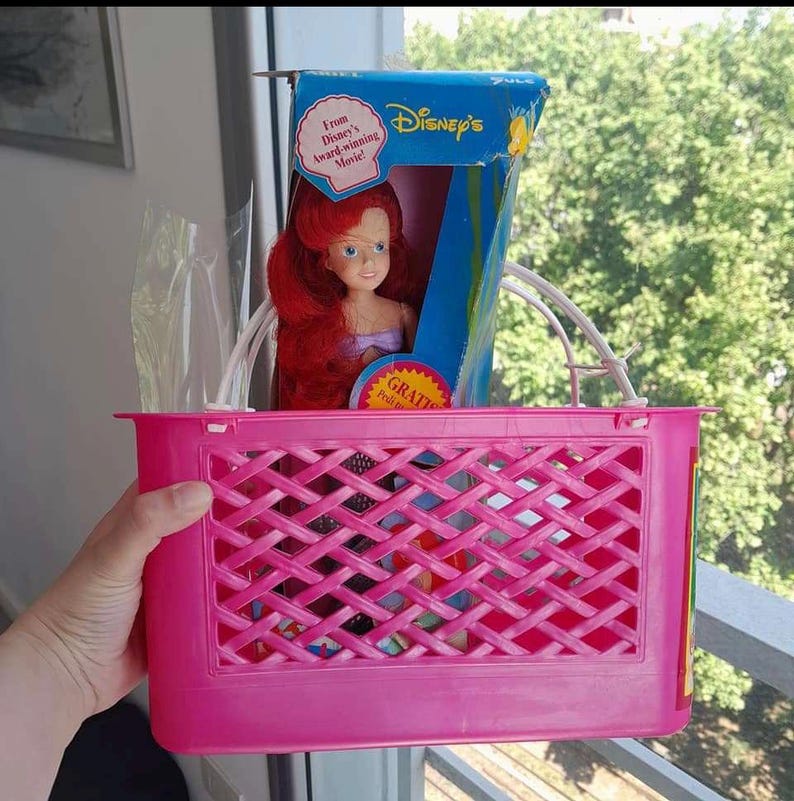 May include: A pink plastic basket with a white handle, containing a red-haired Ariel doll from Disney's The Little Mermaid. The doll is in its original packaging, which features a blue background and the words "From Disney's Award-Winning Movie".