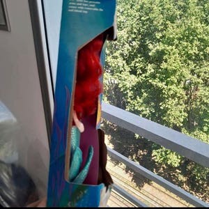 May include: A cardboard box with a blue and green background featuring a mermaid with red hair and a teal tail. The box is partially open and the mermaid doll is visible inside.