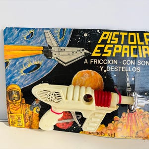 Pode incluir: Pistola espacial de brinquedo vintage com corpo creme e detalhes vermelhos, exibida em uma embalagem de papelão com gráficos espaciais. A embalagem diz "Pistola Espacial".