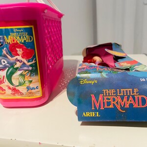 May include: Pink plastic basket with a The Little Mermaid graphic featuring Ariel, Flounder, and Sebastian. The basket is empty. A The Little Mermaid doll packaging is also shown with the text "Disney's The Little Mermaid Ariel" and "Stk No. 1801".