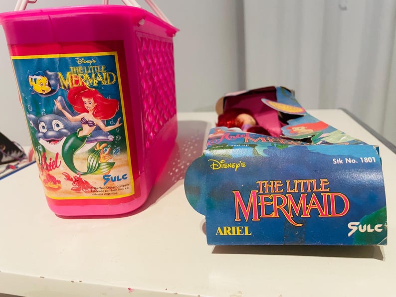 May include: Pink plastic basket with a The Little Mermaid graphic featuring Ariel, Flounder, and Sebastian. The basket is empty. A The Little Mermaid doll packaging is also shown with the text "Disney's The Little Mermaid Ariel" and "Stk No. 1801".