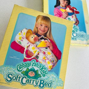 Puede incluir: Dos cajas vintage de Cabbage Patch Kids Soft Carry Bed. Las cajas presentan imágenes de niños con muñecas Cabbage Patch Kids. Las cajas son amarillas con detalles en azul y verde y el texto "Cabbage Patch Kids Soft Carry Bed".