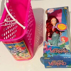 May include: A pink plastic shopping basket with a handle and a picture of Ariel from The Little Mermaid on the front. The basket is next to a doll of Ariel from The Little Mermaid in a blue and green mermaid costume. The doll is packaged in a box with the words "Ariel The Little Mermaid" on it.