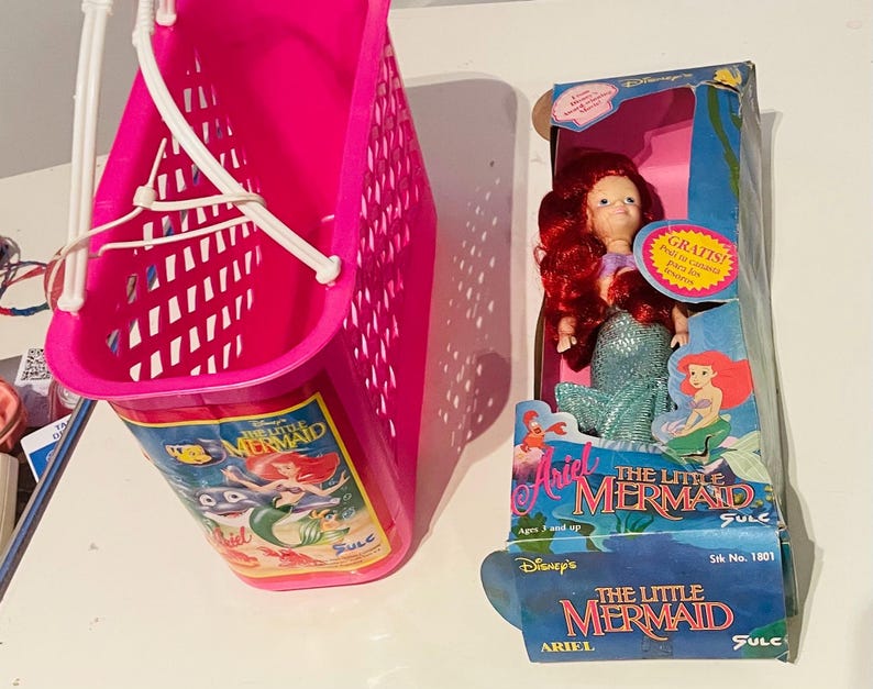 May include: A pink plastic shopping basket with a handle and a picture of Ariel from The Little Mermaid on the front. The basket is next to a doll of Ariel from The Little Mermaid in a blue and green mermaid costume. The doll is packaged in a box with the words "Ariel The Little Mermaid" on it.