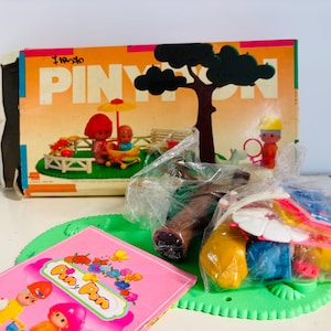 Puede incluir: Un juego de Pinypon vintage con una caja que presenta una escena de parque. El juego incluye figuras, un árbol, un banco y otros accesorios. También se ve un folleto rosa con el logotipo de Pinypon, junto con una base verde y otros juguetes.