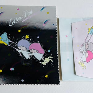 Puede incluir: Un juego de papelería con un bloc de notas y un sobre con un tema celestial. El bloc de notas tiene un fondo negro con estrellas fugaces y personajes de dibujos animados. El juego incluye un sobre azul claro y una tarjeta rosa.