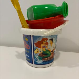 May include: A children's beach toy set featuring a white bucket with a handle, a red bowl, a green bottle, and a yellow shovel. The bucket has a graphic of Ariel and Prince Eric from The Little Mermaid.