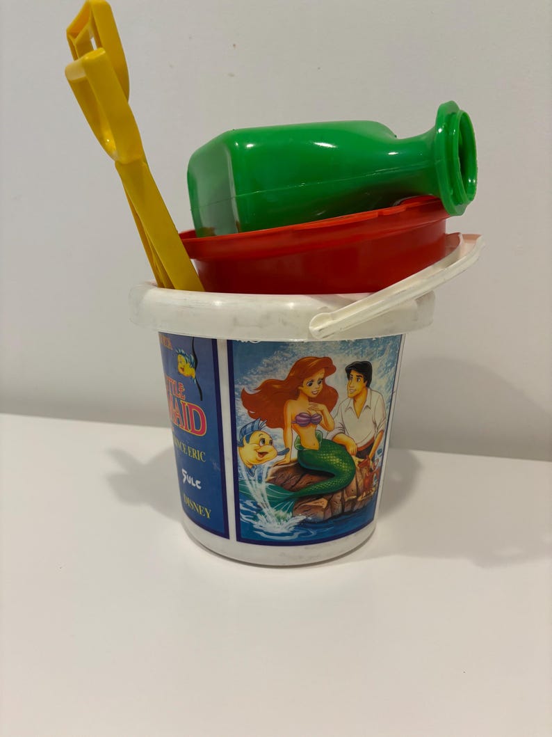 May include: A children's beach toy set featuring a white bucket with a handle, a red bowl, a green bottle, and a yellow shovel. The bucket has a graphic of Ariel and Prince Eric from The Little Mermaid.