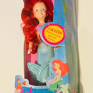 May include: A red-haired Ariel doll from Disney's The Little Mermaid film. The doll is wearing a blue and green mermaid tail and has a pink background. The box also features the text "GRATIS! Pedi tu canasta para los tesoros" and "The Little Mermaid SULC".