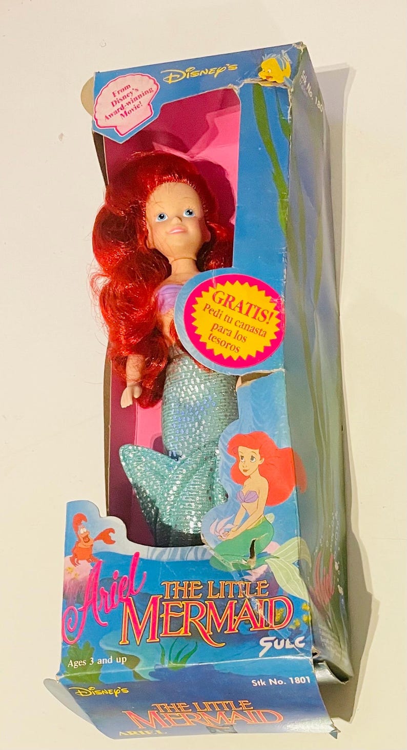 May include: A red-haired Ariel doll from Disney's The Little Mermaid film. The doll is wearing a blue and green mermaid tail and has a pink background. The box also features the text "GRATIS! Pedi tu canasta para los tesoros" and "The Little Mermaid SULC".