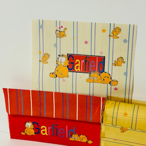Puede incluir: Tres cajas temáticas de Garfield en la imagen. Las cajas son amarillas, rojas y crema, con ilustraciones de Garfield y el nombre "Garfield" impreso. Las cajas tienen patrones de rayas y flores.