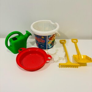 May include: A collection of colorful beach toys. Includes a green watering can, a red sieve, a bucket with "The Little Mermaid" graphics, yellow shovels, a rake, and white sand molds. Perfect for outdoor play.