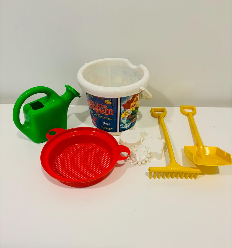May include: A collection of colorful beach toys. Includes a green watering can, a red sieve, a bucket with "The Little Mermaid" graphics, yellow shovels, a rake, and white sand molds. Perfect for outdoor play.