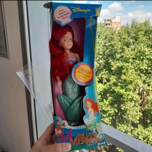 May include: A vintage Disney's The Little Mermaid doll in a box. The doll has red hair and is wearing a green mermaid tail. The box is blue and features a picture of Ariel on the front. The text on the box reads "From Disney's Award-winning Movie!" and "GRATIS! Pedi tu canasta para los fesores".