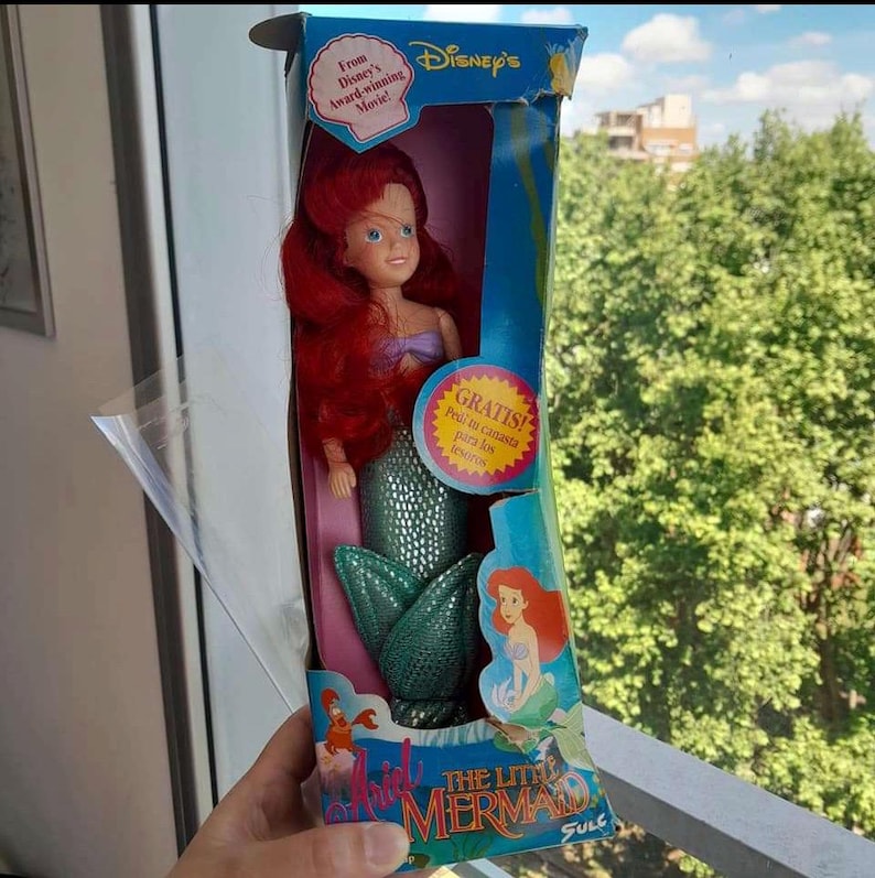 May include: A vintage Disney's The Little Mermaid doll in a box. The doll has red hair and is wearing a green mermaid tail. The box is blue and features a picture of Ariel on the front. The text on the box reads "From Disney's Award-winning Movie!" and "GRATIS! Pedi tu canasta para los fesores".