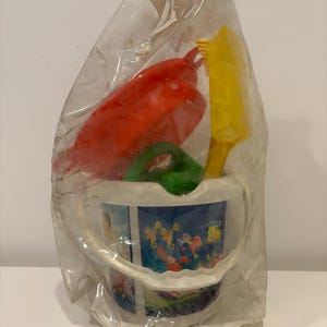 May include: A clear plastic bag containing a children's beach toy set. The set includes a red rake, yellow shovel, green scoop, and a blue bucket with a colorful underwater scene printed on it. The toys are made of plastic.