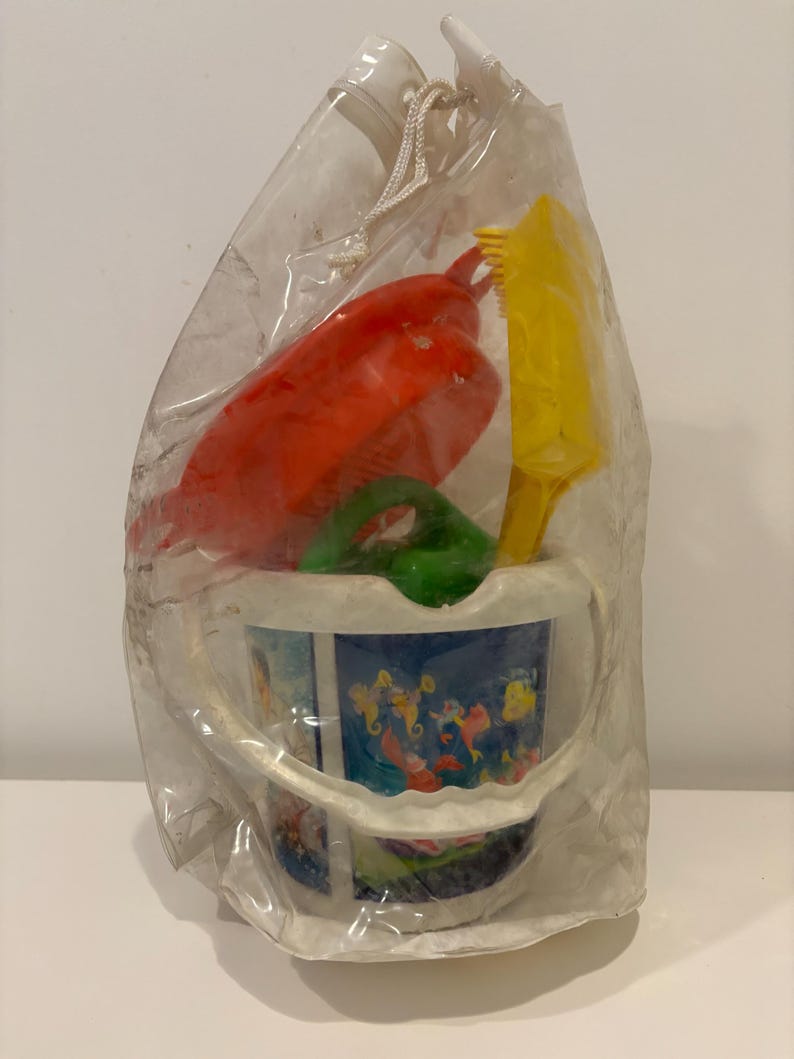 May include: A clear plastic bag containing a children's beach toy set. The set includes a red rake, yellow shovel, green scoop, and a blue bucket with a colorful underwater scene printed on it. The toys are made of plastic.