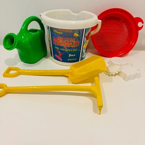 May include: A collection of colorful beach toys, including a green watering can, a white and blue bucket with "The Little Mermaid" graphics, a red sieve, yellow shovels, and white sand molds. Perfect for a day at the beach or sandbox.