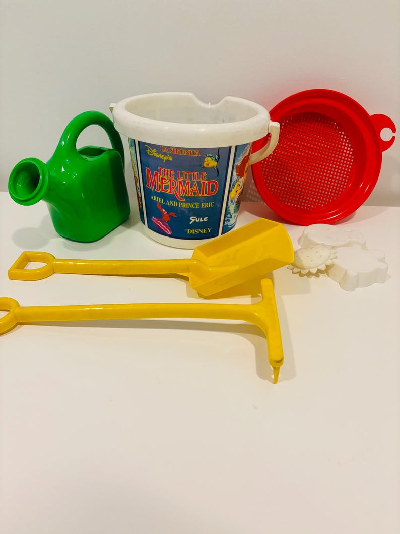 May include: A collection of colorful beach toys, including a green watering can, a white and blue bucket with "The Little Mermaid" graphics, a red sieve, yellow shovels, and white sand molds. Perfect for a day at the beach or sandbox.