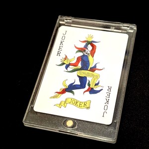 May include: A Joker playing card in a clear plastic case. The card features a colorful illustration of a jester with the word "JOKER" on the top and bottom of the card and on a banner.